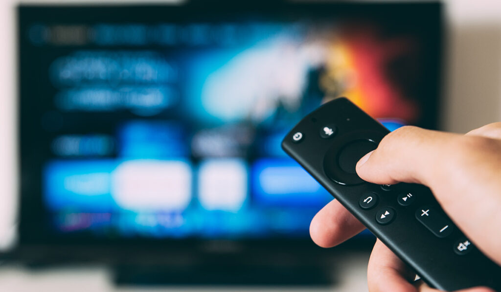 Close up of a hand holding a TV remote against an out of focus TV