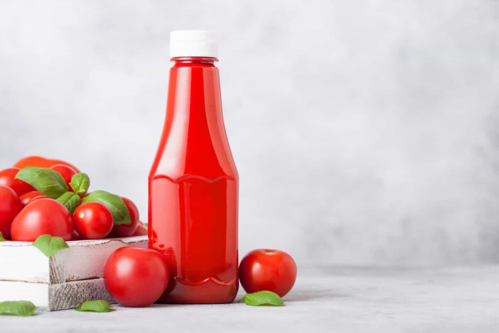 Plastic container with tomato ketchup sauce with raw tomatoes on kitchen background.