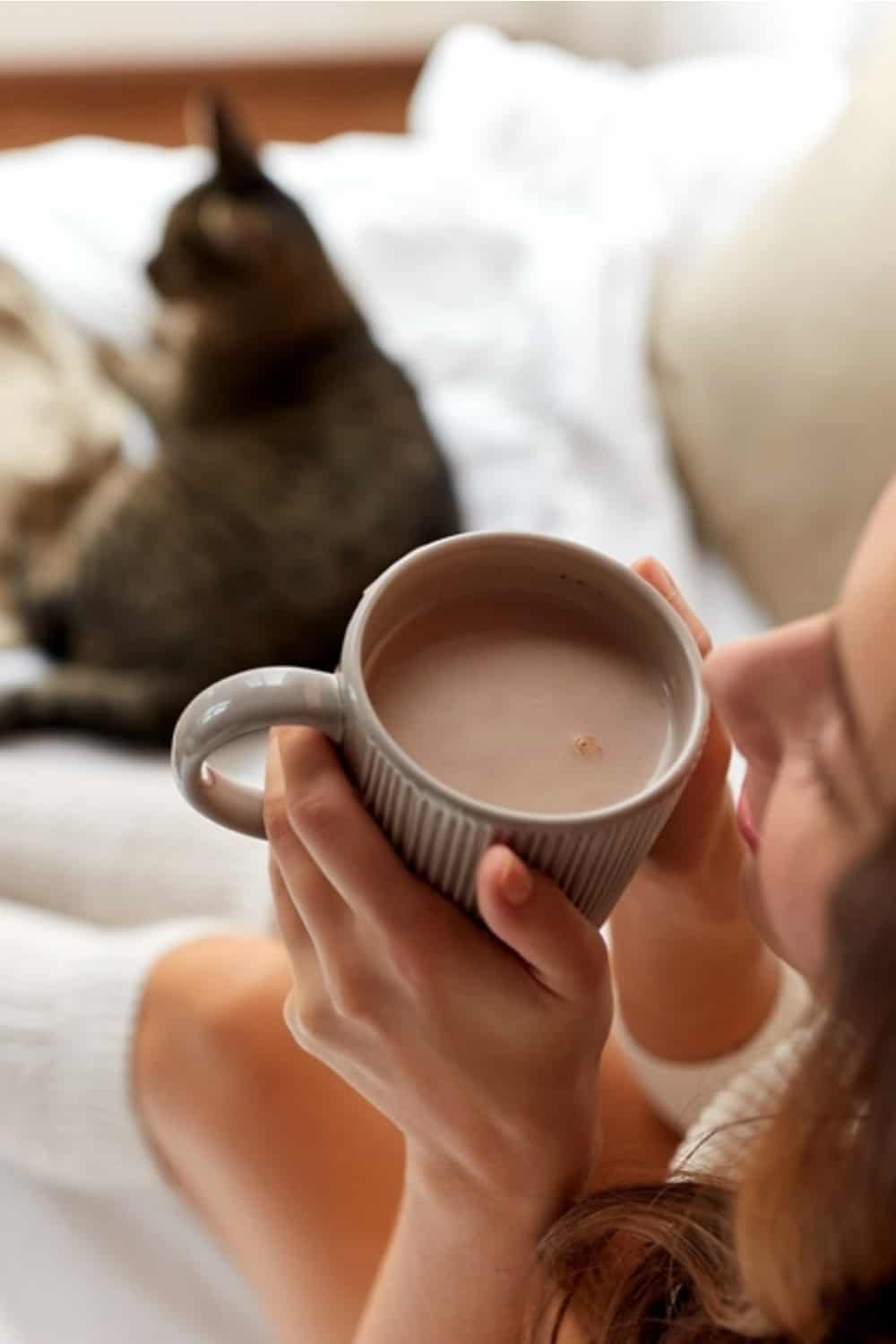 the cat is lying on the bed next to the woman drinking coffee